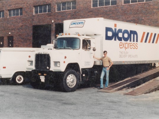 Man standing in front of a Dicom Express truck parked at a terminal in the 1980's